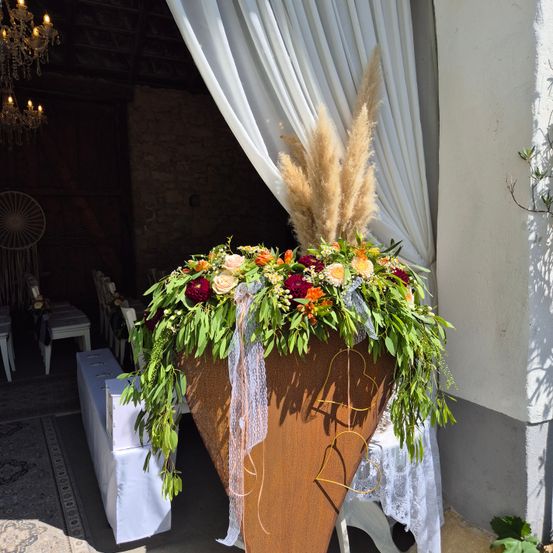 Ein großer brauner Blumentopf voller bunter Blumen und Grünpflanzen steht in der Mitte, mit weißen Vorhängen im Hintergrund und einer sichtbaren Kristallkronleuchter.
