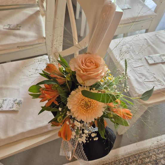 Ein Blumenarrangement mit pfirsichfarbenen Rosen, orangefarbenen Lilien und grünen Blättern ruht auf einer schwarzen Oberfläche, umgeben von weißen Stühlen und Tischdecken.