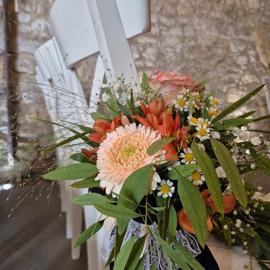 Ein Hochzeitsstrauß mit rosa und orangefarbenen Blumen, gebunden mit einer weißen Spitze, steht auf einem weißen Holzstuhl vor einer Steinmauer.