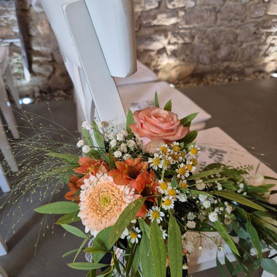 Ein weißer Stuhl, geschmückt mit einem Blumenarrangement aus Rosen, Gänseblümchen und Blättern auf einem Betonboden mit einer Steinmauer im Hintergrund.