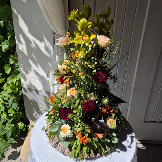 Eine große Blumenarrangement aus Rosen, Lilien und anderen Blumen in leuchtenden Farben steht auf einem Tisch draußen. Das Arrangement steht gegen eine weiße Wand, wobei das Sonnenlicht Schatten wirft.
