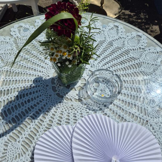 Ein Glastisch mit einem weißen Doily und einer Blumenarrangement. Die Blumen bestehen aus roten, weißen und gelben Blüten. Ein Glasaschenbecher steht auf dem Tisch. Zwei weiße Papierfächer sind auf dem Tisch platziert.