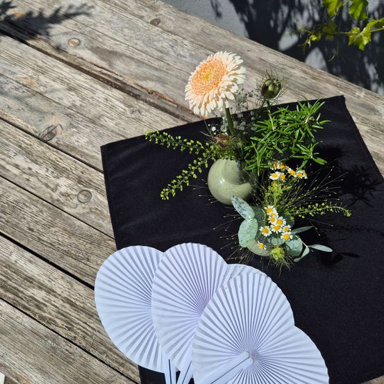 Ein Blumenarrangement mit einer einzelnen weißen Blume und Gänseblümchen liegt auf einem schwarzen Tuch auf einem Holztisch. Links sind zwei weiße Fächer.