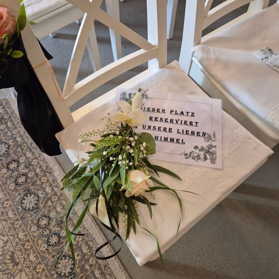Ein weißer Stuhl, geschmückt mit einem Blumenstrauß und einem Schild, das 'Dieser Platz ist reserviert für unsere Lieben im Himmel' steht. Der Stuhl ist mit einem weißen Tuch bedeckt.
