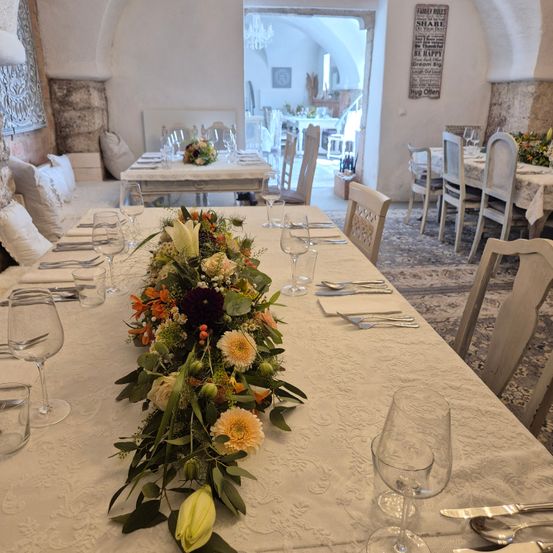 Ein Esszimmer mit einem langen Tisch, der für eine Mahlzeit gedeckt ist. Der Tisch hat eine weiße Tischdecke und einen Blumen-Zentren. Weingläser und Besteck sind auf dem Tisch arrangiert. Andere Tische und Stühle sind im Hintergrund sichtbar.