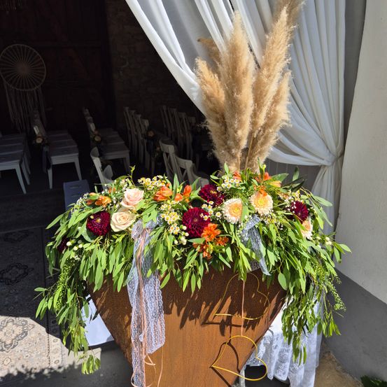 Ein großes Blumenarrangement in einem braunen Pflanzgefäß steht vor einem weißen Vorhang, bestehend aus einer Mischung aus roten, orangefarbenen und weißen Blumen und dekorativem Band.