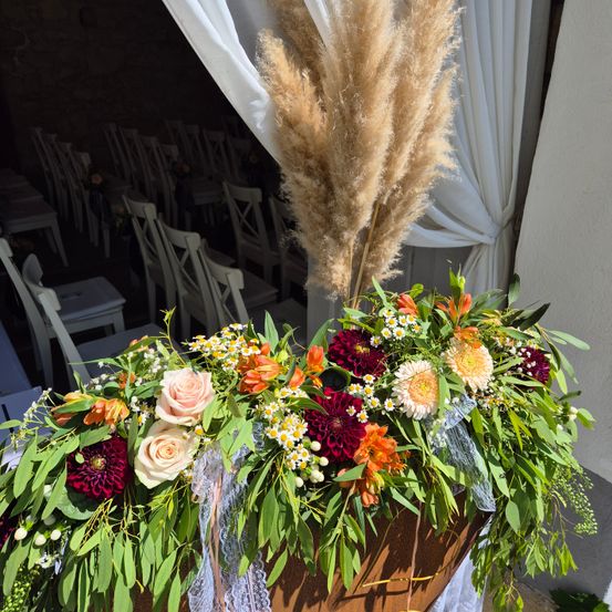 Ein Hochzeitsgang mit Stühlen ist mit einer großen Blumenarrangement dekoriert. Das Zentrum umfasst Rosen, Gänseblümchen und Herbstblumen, geschmückt mit Pampasgras.