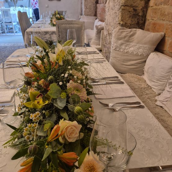 Ein langer Esstisch mit einem Blumenarrangement in der Mitte und verschiedenen Tischutensilien, darunter Gläser und Besteck, die für eine Mahlzeit vorbereitet sind. Dahinter eine Ziegelwand und eine Couch mit Kissen.