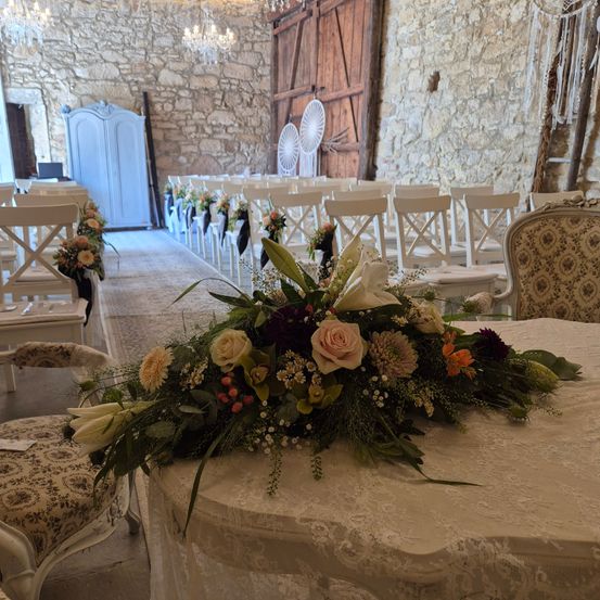 Eine Hochzeitszeremonie mit Steinmauer, Holztüren und weißen Stühlen. Ein Blumenarrangement steht auf dem Tisch.