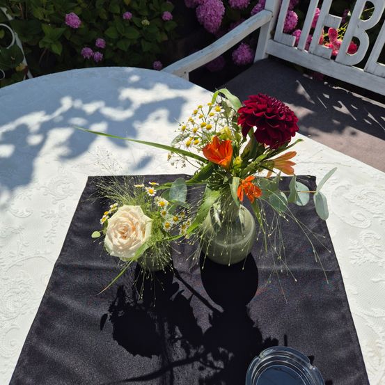 Ein Tisch mit einer weißen Tischdecke hat zwei Vasen mit Blumen auf einem schwarzen Tischläufer. Eine Vase hat eine Rose und andere Blumen, während die andere rote und orange Blumen hat.