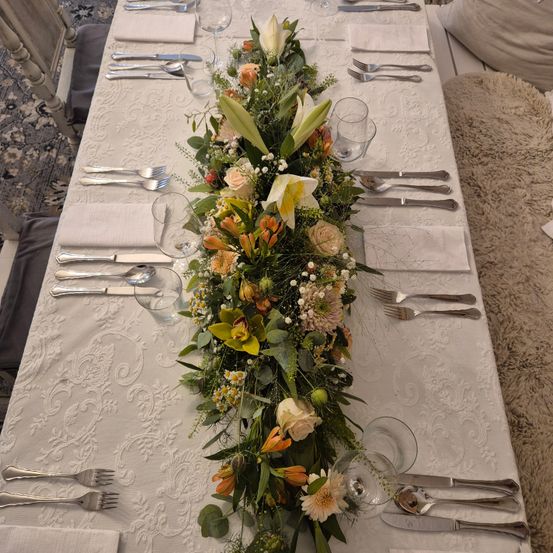 Ein langer Tisch für eine formelle Veranstaltung mit Blumen als Mittelpunkt, Silberbesteck und Servietten. Der Tisch ist mit einer weißen Tischdecke bedeckt.