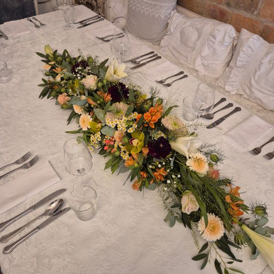 Ein langes Blumenarrangement mit verschiedenen Blumen wie Rosen, Lilien und Gänseblümchen, auf einem weißen Tischtuch mit Besteck und Servietten.