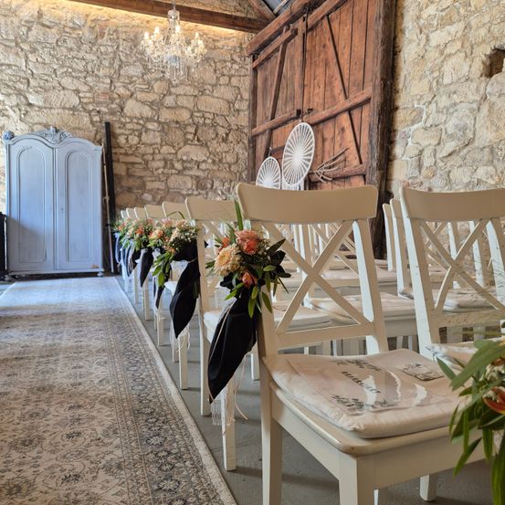 Gang aus weißen Stühlen mit Blumenarrangements und schwarzen Schärpen. Steinwände und Holztür im Hintergrund. Kronleuchter und Schrank im Hintergrund.