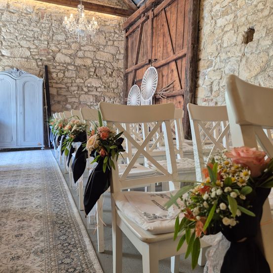 Eine Reihe weißer Stühle, geschmückt mit Blumen und schwarzem Stoff, steht vor Steinmauern und einer Holztür.