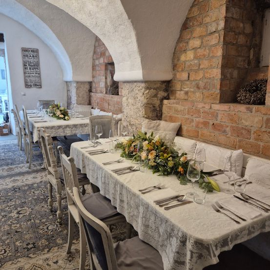 Ein Essbereich mit zwei Tischen, beide zum Essen gedeckt mit Blumen, Besteck und Gläsern. Der Raum hat Backsteinwände und einen Teppich auf dem Boden.
