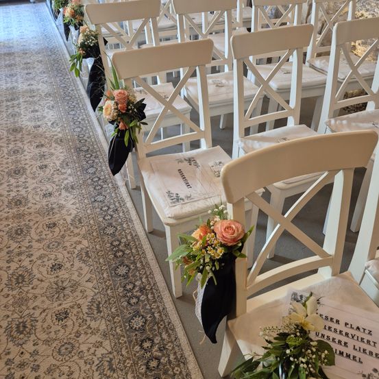 Gang mit weißen Stühlen und Blumenarrangements, jeder Stuhl hat ein schwarzes Band mit Blumen auf der Rückseite. Ein Teppichboden und ein Tisch mit Blumen und einem Schild.