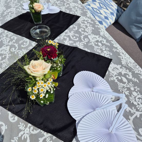 Ein Tisch ist mit schwarzen Platzdeckchen und weißen dekorativen Fächern gedeckt. Es gibt Blumenarrangements in Glasvasen auf dem Tisch. Eine Anordnung umfasst eine Rose, Gänseblümchen und andere Blumen. Eine weitere Anordnung befindet sich im Hintergrund mit grünen Blättern. Der Tisch hat eine weiß-grau gemusterte Tischdecke.