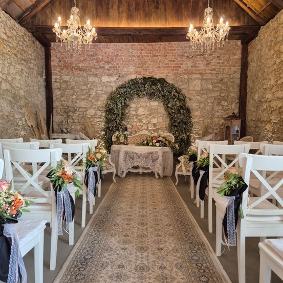 Ein Hochzeitszeremoniebereich mit weißen Stühlen, einer Steinmauer und einem Tisch mit Blumen. Ein dekorativer Bogen aus Grünpflanzen führt zum Altar.