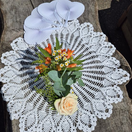Ein weißer gehäkeltes Doily mit einer Blumenarrangement aus orange und weißen Blumen, grünen Blättern und einer weißen Rose auf einer Holzoberfläche.