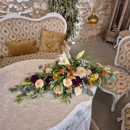 Ein üppiges Blumenarrangement ziert einen Tisch in einer rustikalen Umgebung. Der Tisch ist mit Spitze bedeckt, und ein Sofa mit verzierten Kissen steht in der Nähe.