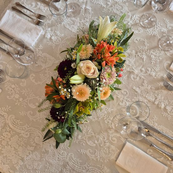 Ein Tisch für eine formelle Veranstaltung mit Blumenarrangement, weißer Tischdecke, Besteck und Weingläsern. Das Blumenarrangement besteht aus weißen, orangefarbenen und gelben Blumen.