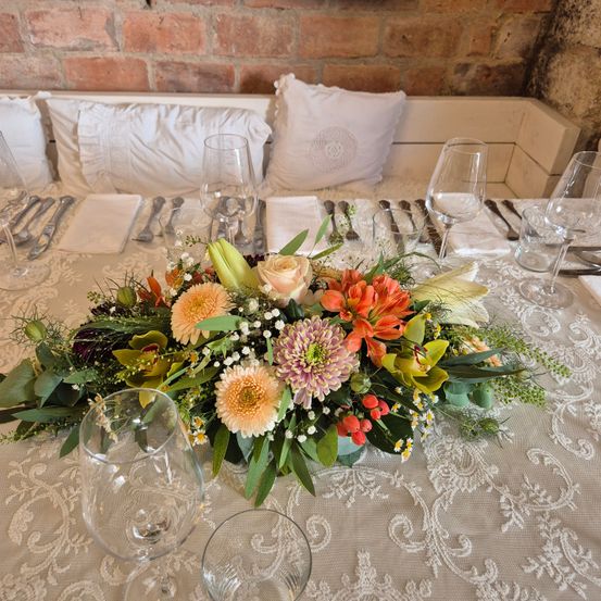 Ein Tisch ist mit Weingläsern, Besteck und einem Blumencenterpiece dekoriert. Der Tisch hat eine weiße Tischdecke mit einem komplizierten Design. Hinter dem Tisch befindet sich eine Ziegelwand mit einer Bank und Kissen.