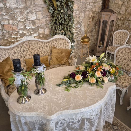 Ein Raum mit einem Sofa und einem Tisch, beide mit Spitze bedeckt, hat zwei Kerzen und einen Blumenstrauß. Der Raum hat eine Steinmauer und zwei Stühle sind in der Nähe platziert.