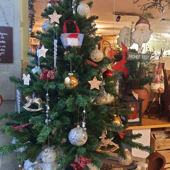 Ein mit Ornamenten und Dekorationen geschmückter Weihnachtsbaum, einschließlich einer Santa-Claus-Figur, hängt in einem Geschäft. Hinter dem Baum steht ein Holztresen mit verschiedenen Artikeln.