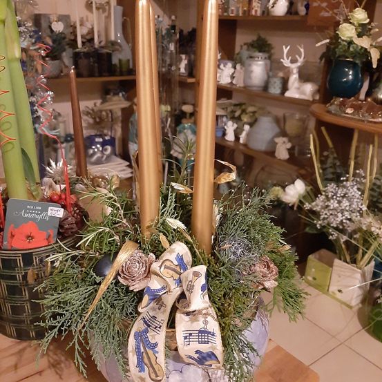 Eine dekorative Anordnung mit zwei hohen Kerzen, umgeben von Grün und einem Band, ausgestellt in einem Laden mit Regalen verschiedener Gegenstände im Hintergrund.