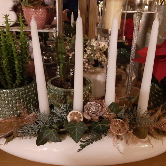 Eine dekorative Anordnung mit vier weißen Kerzen, geschmückt mit Grün, getrockneten Blumen und Zitrusscheiben, die auf einer weißen ovalen Basis auf einem Holztisch ruht.