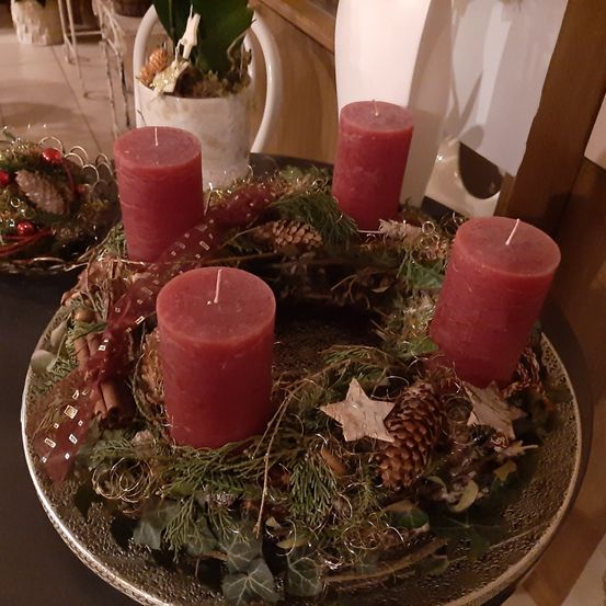 Ein Kranz mit roten Kerzen, Tannenzapfen und Stechpalme wird auf einem Tisch ausgestellt. Die Kerzen sind nicht angezündet. Dahinter steht eine Pflanze in einem weißen Topf.