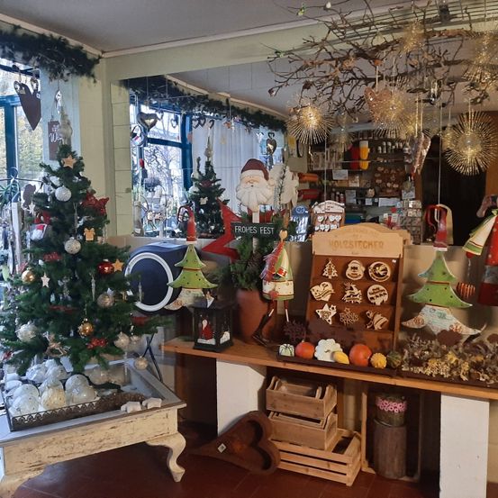 Ein Weihnachtsbaumgeschäft mit verschiedenen Dekorationen und Ornamenten. Eine Santa-Statue und ein Weihnachtsbaum sind rechts platziert. Ein hölzerner Schreibtisch mit Kürbissen, Holzkisten und Blumenvasen steht in der Mitte.