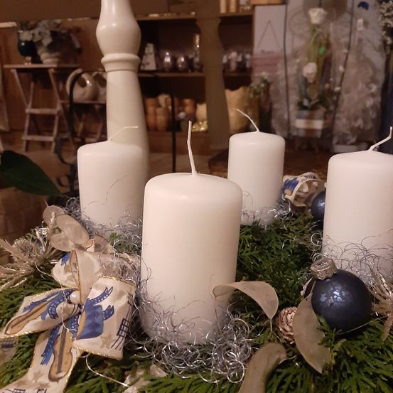 Vier weiße Kerzen sind in einem Kranz mit Grün, Silbertinsel und Bändern angeordnet. Ein blau-weißes Band hat eine Schleife. Es gibt einen dunkelblauen Ornament.