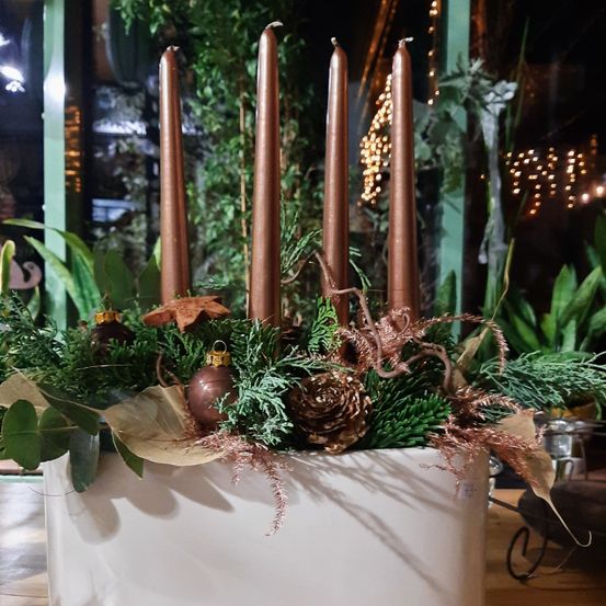 Ein weißer Pflanzkasten enthält eine festliche Dekoration mit vier hohen braunen Kerzen, umgeben von Grün, Tannenzapfen und Ornamenten, auf einer Holzoberfläche platziert.