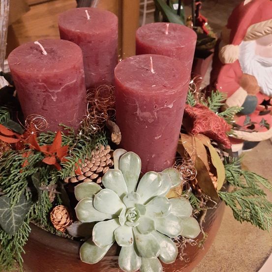 In einer Holzschale befinden sich vier rote Kerzen, umgeben von Tannenzapfen, Blättern und einer Sukkulente. Im Hintergrund steht eine Weihnachtsmannfigur.