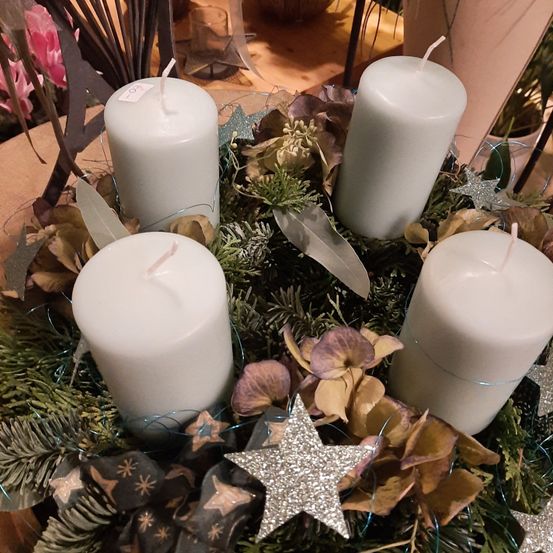 Vier weiße Kerzen sind in einem Kranz mit Silbersternen, Grünpflanzen und getrockneten Blumen auf einem Tisch angeordnet.