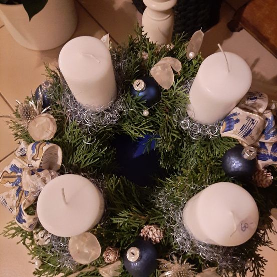 Ein Kranz mit blauen und silbernen Dekorationen, bestehend aus vier weißen Kerzen, steht auf einer gefliesten Oberfläche. Der Kranz ist mit Tannenzapfen, Perlen und einer blauen Kugel verziert.