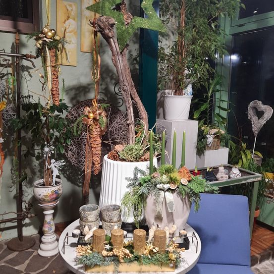 Ein Raum mit mehreren Pflanzen, Vasen und Dekorationsartikeln. Im Mittelpunkt steht eine weiße Vase mit einer Schleife, umgeben von grünen Pflanzen und Kerzen. Dahinter befindet sich ein großer Ast und verschiedene andere Dekorationselemente.