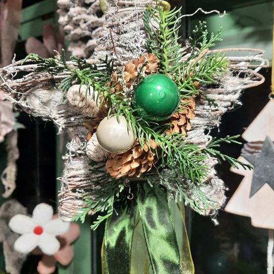 Ein sternförmiger Weihnachtsschmuck mit einem grünen Band. Er hat einen grünen Ball, einen weißen Ball und Tannenzapfen. Der Stern ist aus grünen Blättern und Zweigen gefertigt und hängt vor einem grünen Hintergrund.