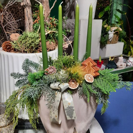 Eine dekorative Komposition mit vier grünen Kerzen in einem rosafarbenen Topf, umgeben von Grünpflanzen, einer Schleife und getrockneten Zitrusscheiben. Im Hintergrund befinden sich Topfpflanzen und weitere Dekorationen.