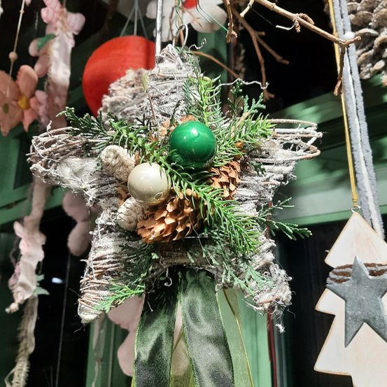 Ein sternförmiger Schmuck mit grünen Zweigen und einer grünen Kugel in der Mitte, der an einem Ast hängt und von anderen Ornamenten und Blumen umgeben ist.