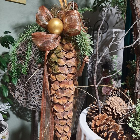 Ein dekorativer Kiefernzapfen mit goldener Kugel und brauner Schleife, umgeben von grünen Blättern und Zweigen, auf einem weißen Topf platziert.