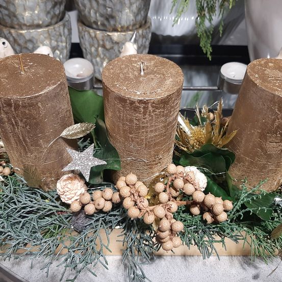 Drei goldene Kerzen sind in einer festlichen Dekoration mit Elementen wie Blättern, Beeren und einem silbernen Stern auf einem Holztablett platziert.