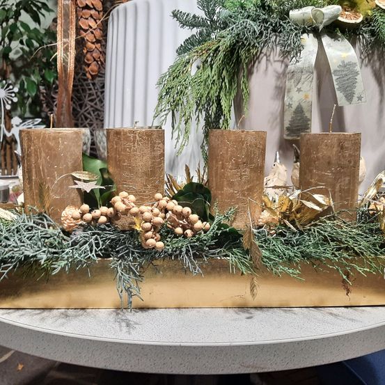 Ein goldenes Tablett hält fünf Kerzen, umgeben von Tannenzweigen, Beeren und goldenen Blättern. Die Kerzen sind mit Glitzer verziert. Ein Kranz und ein Band hängen dahinter.