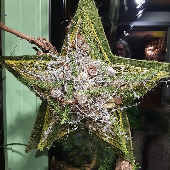Eine sternförmige Dekoration aus grünem Moos und Zweigen wird an einer grünen Wand ausgestellt. Sie hat eine Mischung aus Muscheln und Tannenzapfen darauf. Eine Person steht hinter der Dekoration.