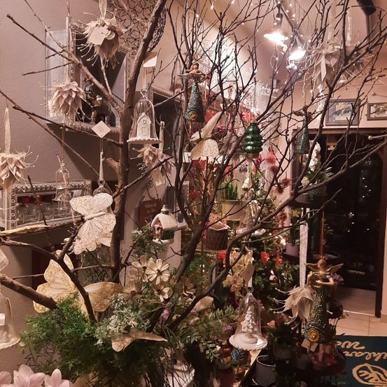 Ein Raum, der mit einer großen baumartigen Struktur verziert ist, die mit verschiedenen Ornamenten und Feenfiguren geschmückt ist, umgeben von Blumen und Lichtern. Regale mit dekorativen Gegenständen und ein Teppichboden.