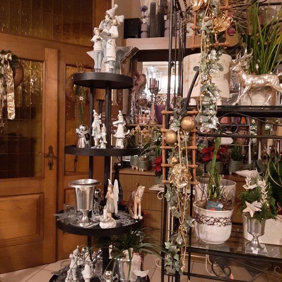 Ein Raum mit einer Ausstellung von Dekorationsartikeln, darunter Figuren, Ornamente und Pflanzen. Ein dreistufiger Ständer mit silbernen Objekten, Figuren und Ornamenten steht in der Mitte. Regale mit Pflanzen und Dekorationsartikeln befinden sich auf beiden Seiten. Eine Holztür und eine Glastür sind im Hintergrund.