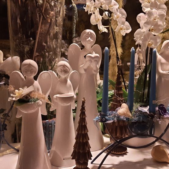 Ein Tisch zeigt Keramikengel, Kerzen und einen kleinen Baum. Jeder Engel hält eine Blume und einen Becher. Dahinter stehen weiße Orchideen in einem Vasen.