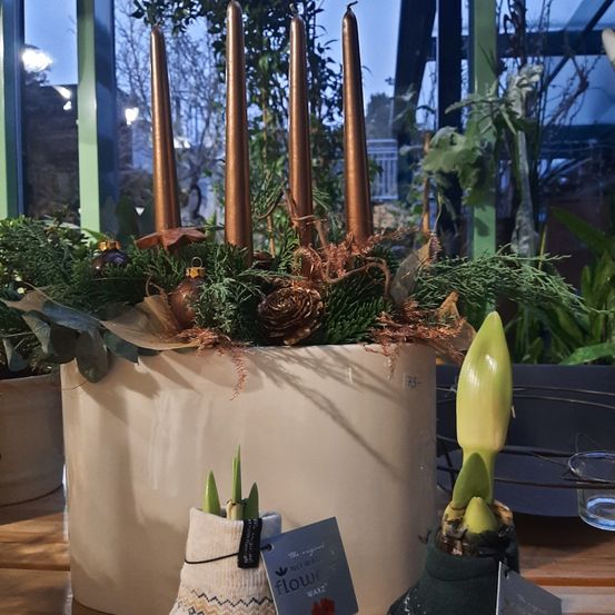 Ein großer beigefarbener Pflanzkübel mit Bronzekerzen und dekorativen Pflanzen. Der Kübel steht auf einem Holztisch mit einem Schild an der Seite. Davor befinden sich zwei kleine Topfpflanzen mit Etiketten.