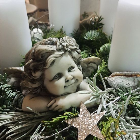 Eine lächelnde Engelsfigur ruht auf einem Bett aus Tannennadeln, umgeben von weißen Kerzen und einem goldenen Stern, möglicherweise für eine festliche Dekoration.